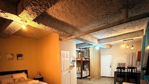 Ein gemütlicher, rustikaler Raum mit einem Bett, einem kleinen Tisch und einer Bank. Ein Durchgang führt in einen weiteren Raum mit einem Bett und einem Spiegel.