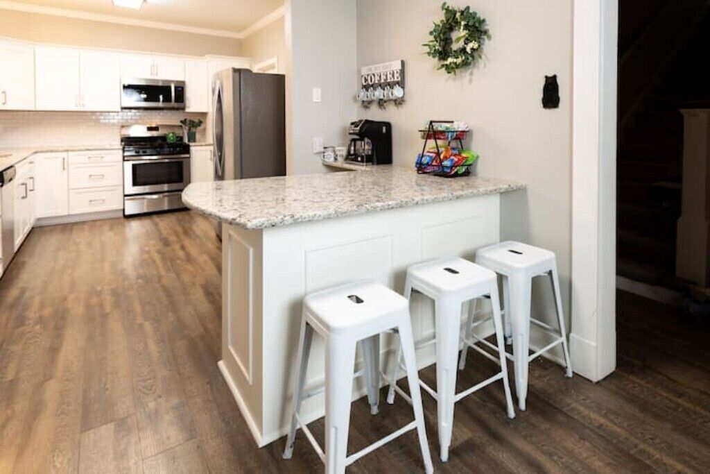 Una cucina moderna con mobili bianchi, elettrodomestici in acciaio inossidabile e un piano di lavoro in granito. Sono presenti tre sgabelli da bar bianchi e un angolo caffè con una macchina per il caffè e un cestino di snack.