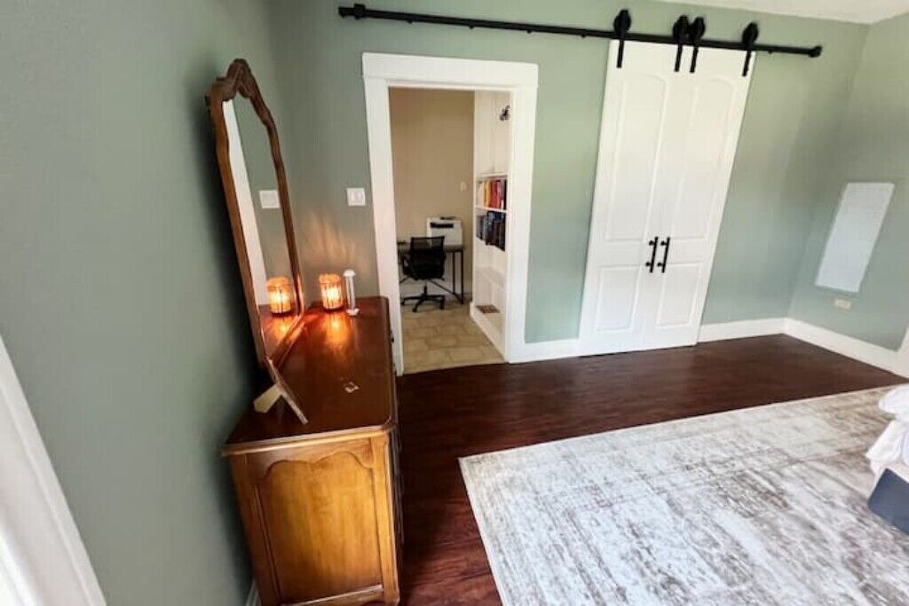 Un dormitorio con un aparador de madera, un espejo y una alfombra blanca. Hay una puerta que da a un pasillo con un perchero y un armario blanco.