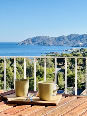 En træterrasse med to keramikkrus og en ske, med udsigt over et kystlandskab med bjerge og grønt.