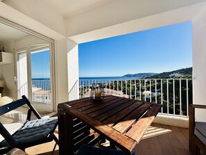 En balkon med et træbord og stole, en glasdør der fører til et indendørs område, og udsigt til hav og bakker.