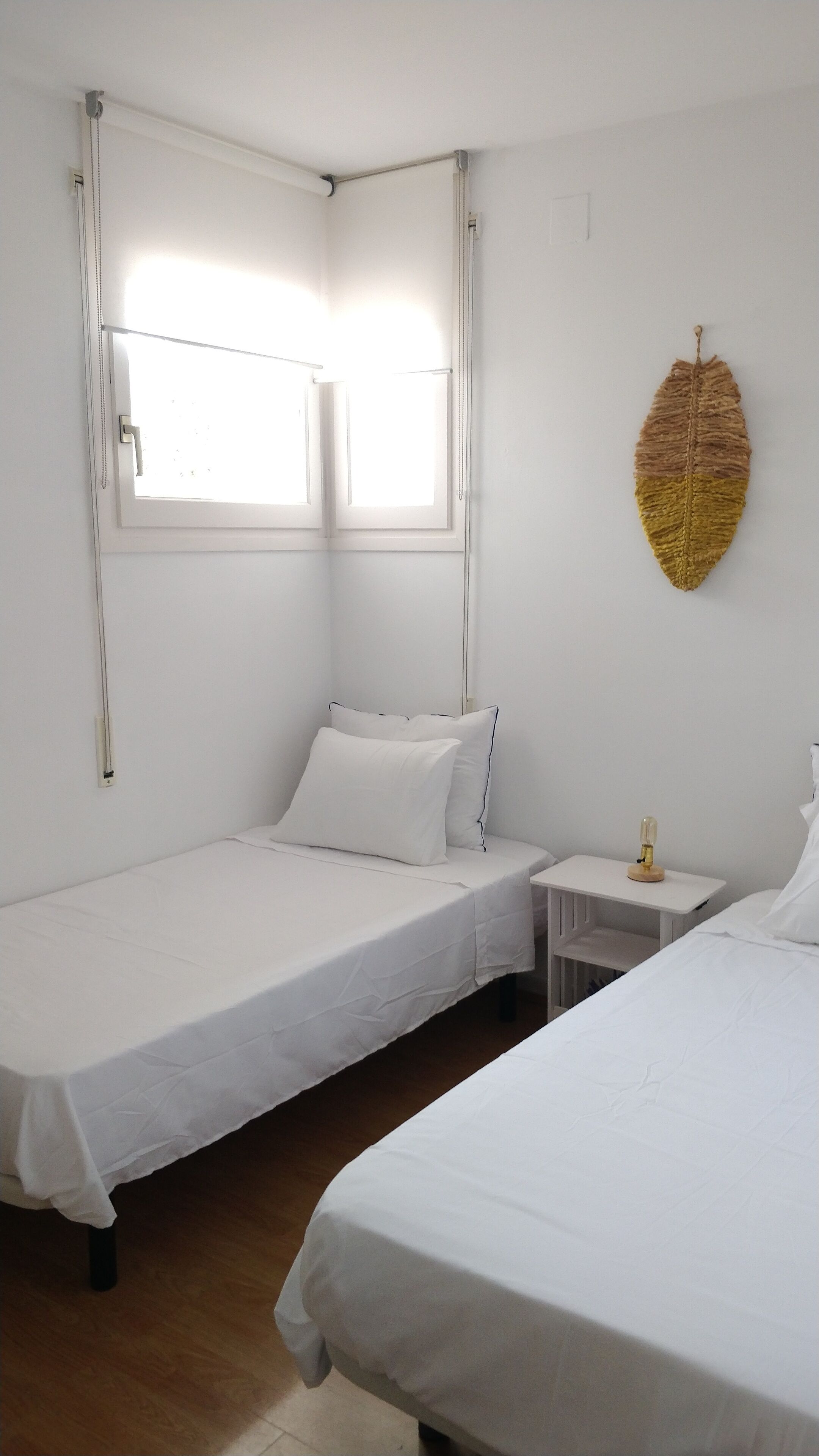 A hotel room with two single beds, a small bedside table with a lamp, a window with blinds, and a wall-mounted decorative item.