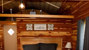 A wooden cabin room with a bed, a nightstand with a lamp, and a wall-mounted shelf with decorative items.