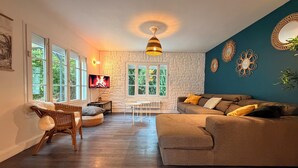 A cozy living room with a stone wall, a large window, a sofa, a TV, and a hanging light fixture. - Les Sablons - Villa 15pers - table football darts bbq (La Houssaye-en-Brie)