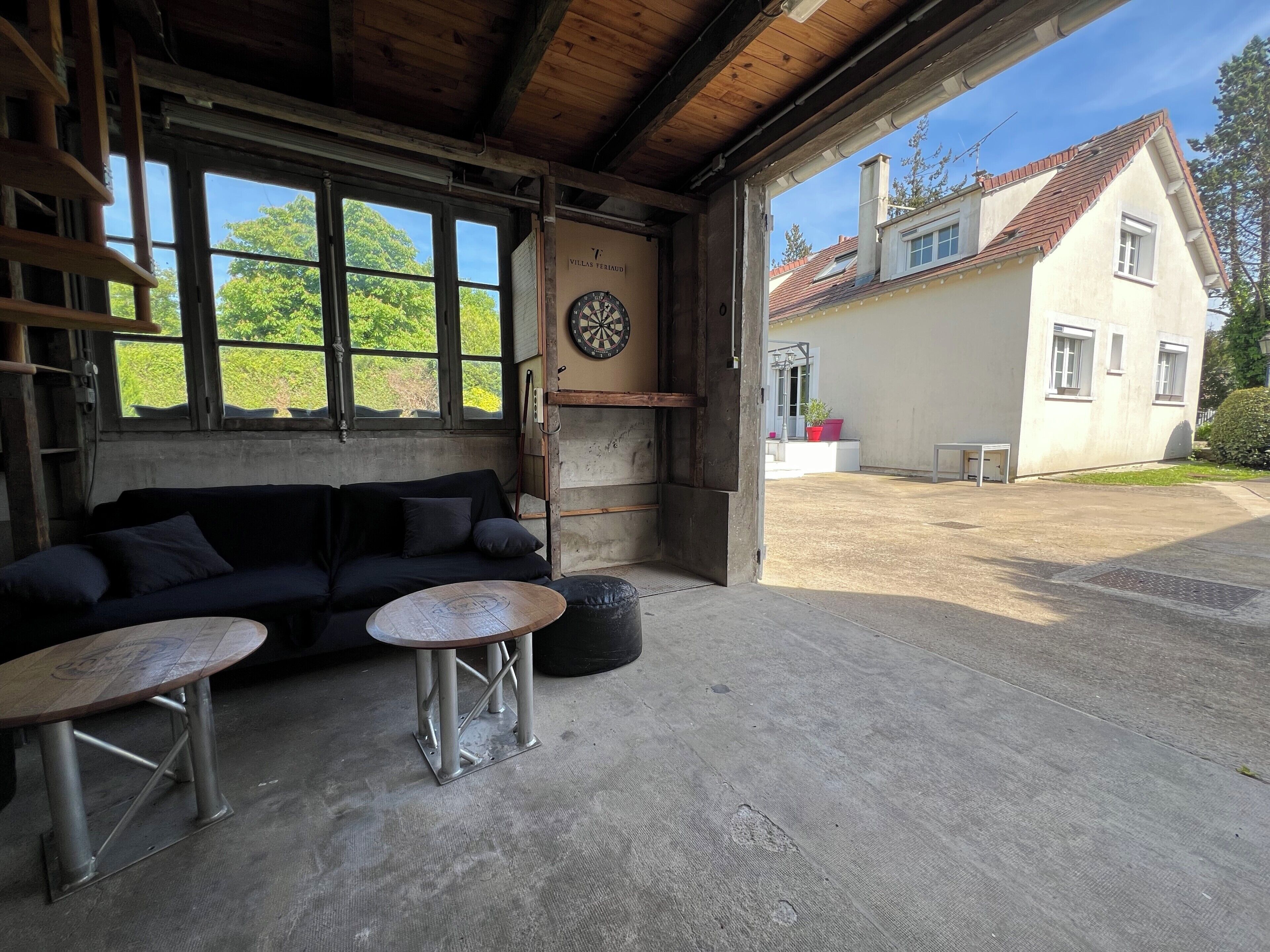 A rustic interior with a wooden ceiling, a black sofa, two metal tables, and a dartboard.