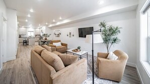 Ein modernes Wohnzimmer mit einem großen, beigefarbenen Ecksofa, einem Couchtisch und einer Stehlampe. Im Hintergrund ist ein Essbereich mit einer sichtbaren Küche zu sehen.