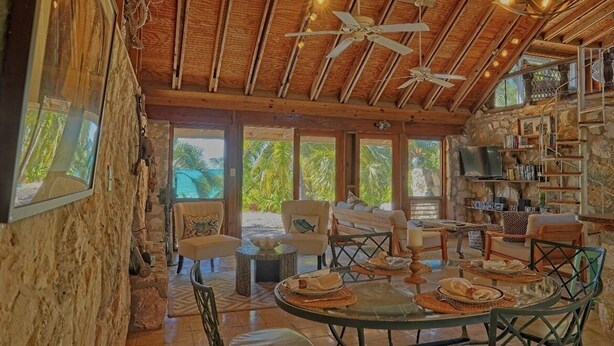 A spacious room with a high ceiling, wooden beams, a stone wall, a large window, and a dining area with a table set for four. - Hibiscus House Oceanfront Paradise in Fernandez Bay (New Bight)