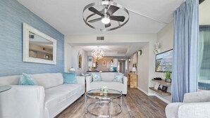 Un salon moderne avec un canapé blanc, une table basse en verre, un ventilateur de plafond et une cuisine en arrière-plan.