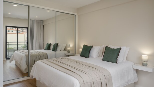A bedroom with a large mirror, a bed with white bedding and green pillows, a bedside table with a lamp, and a view through a glass door. - In the Funchal city center, Santa Luzia III (Funchal)