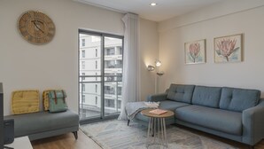 A modern living room with a grey sofa, a small round table, a large window with a view of buildings, and a wall clock. - In the Funchal city center, Santa Luzia III (Funchal)