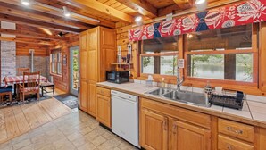 A wooden cabin-style kitchen with a dishwasher, a sink, and a window overlooking a natural landscape.