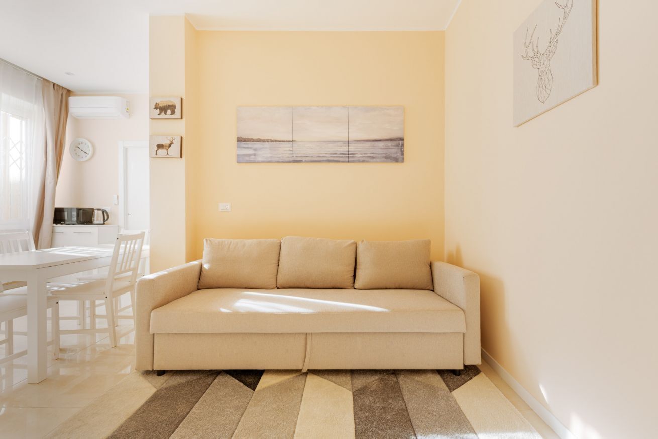 A living room with a beige sofa, a dining table with chairs, a kitchen area, and a wall-mounted artwork.
