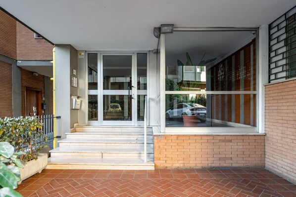 Entrada de un edificio con puertas de vidrio, pared de ladrillo y patio con baldosas.