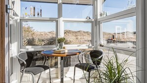 Une pièce lumineuse, baignée de soleil, avec de grandes fenêtres, deux chaises, une petite table et une plante en pot.