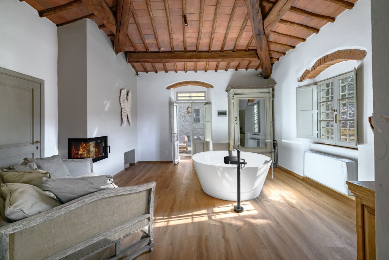 A spacious room with a wooden ceiling, a large white bathtub, a fireplace, and a view of the outside through an arched window.