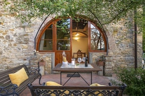 An outdoor seating area with a stone wall, a wooden bench, a table with a bottle, glasses, and a pitcher.