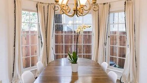 Une salle à manger avec une table en bois, des chaises blanches, un lustre et de grandes fenêtres avec des rideaux.