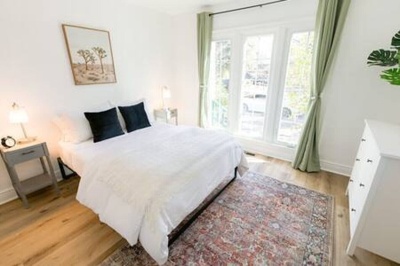 A bedroom with a bed, bedside tables, a nightstand, a window with curtains, and a plant.