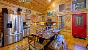 A wooden cabin interior with a dining table, chairs, a red door, and a kitchen with appliances. - Oar & Arrow · Wilderness Retreat: Hot Tub, Grill, King Suites (Broken Bow)