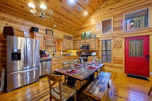 A wooden cabin interior with a dining table, chairs, a red door, and a kitchen with appliances. - Oar & Arrow · Wilderness Retreat: Hot Tub, Grill, King Suites (Broken Bow)