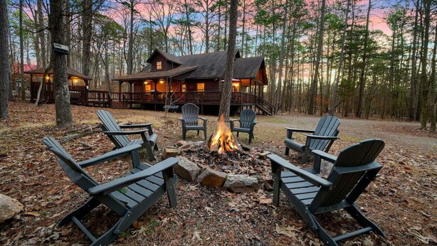A cabin with a fire pit surrounded by Adirondack chairs, set in a forested area. - Oar & Arrow · Wilderness Retreat: Hot Tub, Grill, King Suites (Broken Bow)