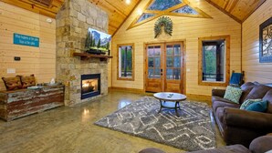 A cabin interior with a stone fireplace, wooden walls, a cozy seating area, and a large window. - The Copper Hideout · Hochatown Hideout: Hot Tub, Fire Pit, Pure Relaxation (Broken Bow)