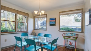 A dining area with a glass table and turquoise chairs, large windows with blinds, and a colorful table lamp.