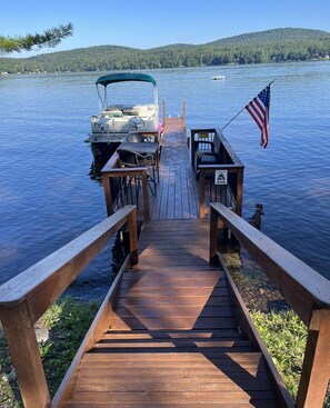 A wooden dock leading to a boat with an American flag, surrounded by calm water and a forested hill in the background. - On Caroga Time - Year Round Cottage With Private Dock (Caroga Lake)