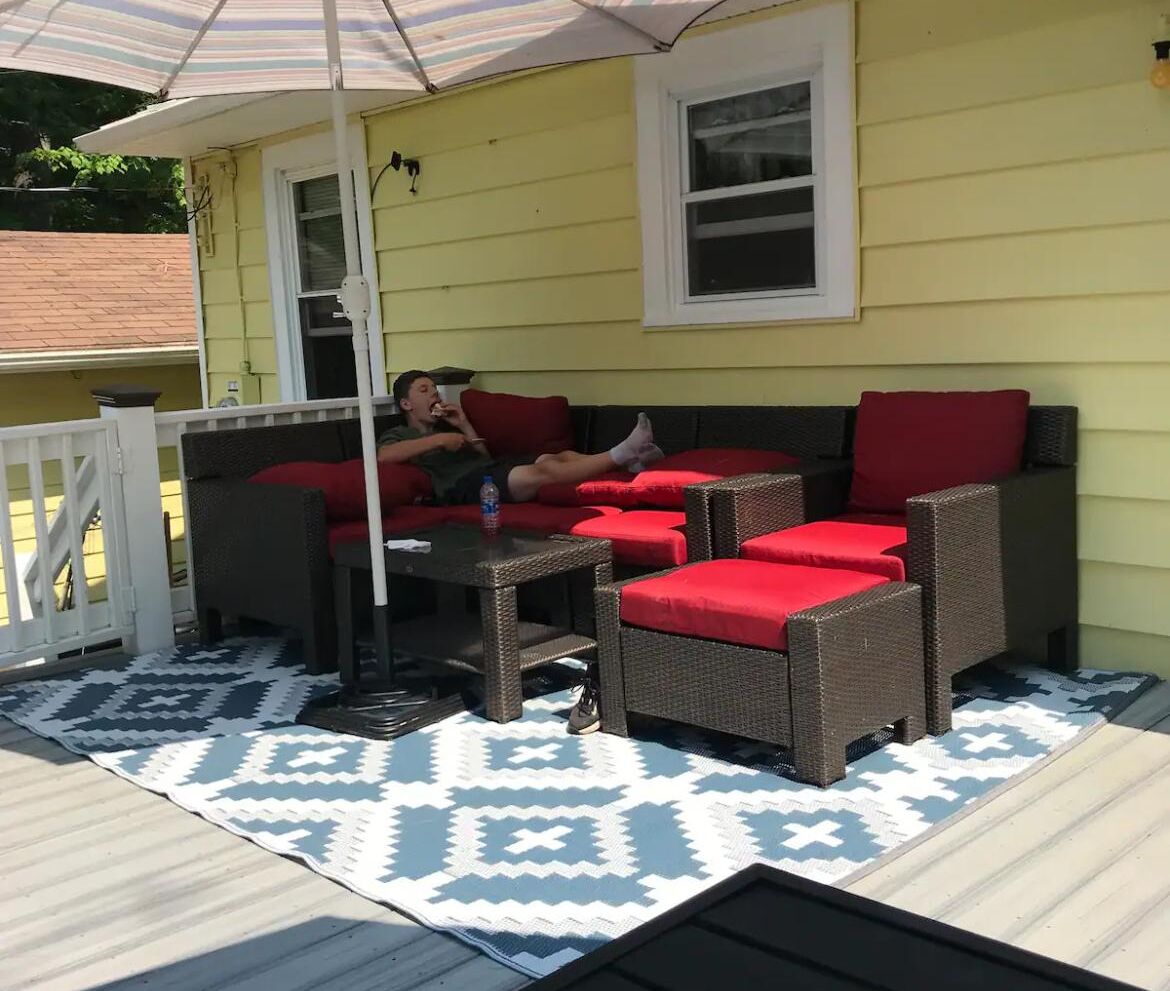 An outdoor patio with wicker furniture and red cushions, a white railing, and a patterned rug.