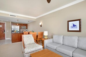 Ein Wohnzimmer mit grauem Sofa, einem floralen Sessel und einem Holztisch. Im Hintergrund ist eine Küche mit Holzschränken und einer Mikrowelle zu sehen.