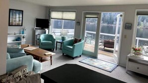 Un salón con sofá, sillones, una mesa de centro y televisor. Hay ventanas amplias y una puerta corrediza que da a un balcón.