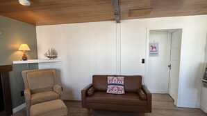 Een woonkamer met een bruine leren bank, een beige fauteuil, een klein tafeltje met een scheepsmodel en een bordje aan de muur.