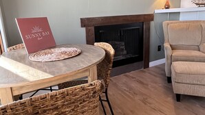 Een gezellige woonkamer met een open haard, een ronde houten tafel met placemat, rieten stoelen en een beige fauteuil.