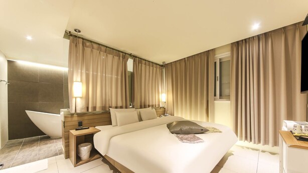 A modern hotel room with a large bed, a bedside table, a nightstand, a bathroom with a bathtub, and a window with curtains.