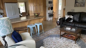 A modern living room with a white sofa, a coffee table, and a black leather couch. There is a kitchen area with wooden cabinets and a refrigerator.