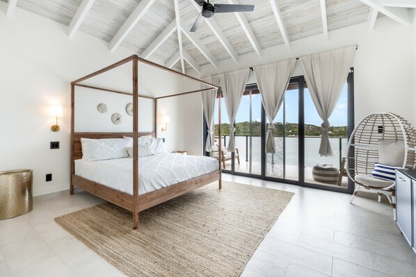 A four-poster bed with a wooden frame, a woven rug, and a view of the sea through large glass doors. - Márohu, 3BR Beach Oasis on Flamingo Lake (Venetian Road Settlement)