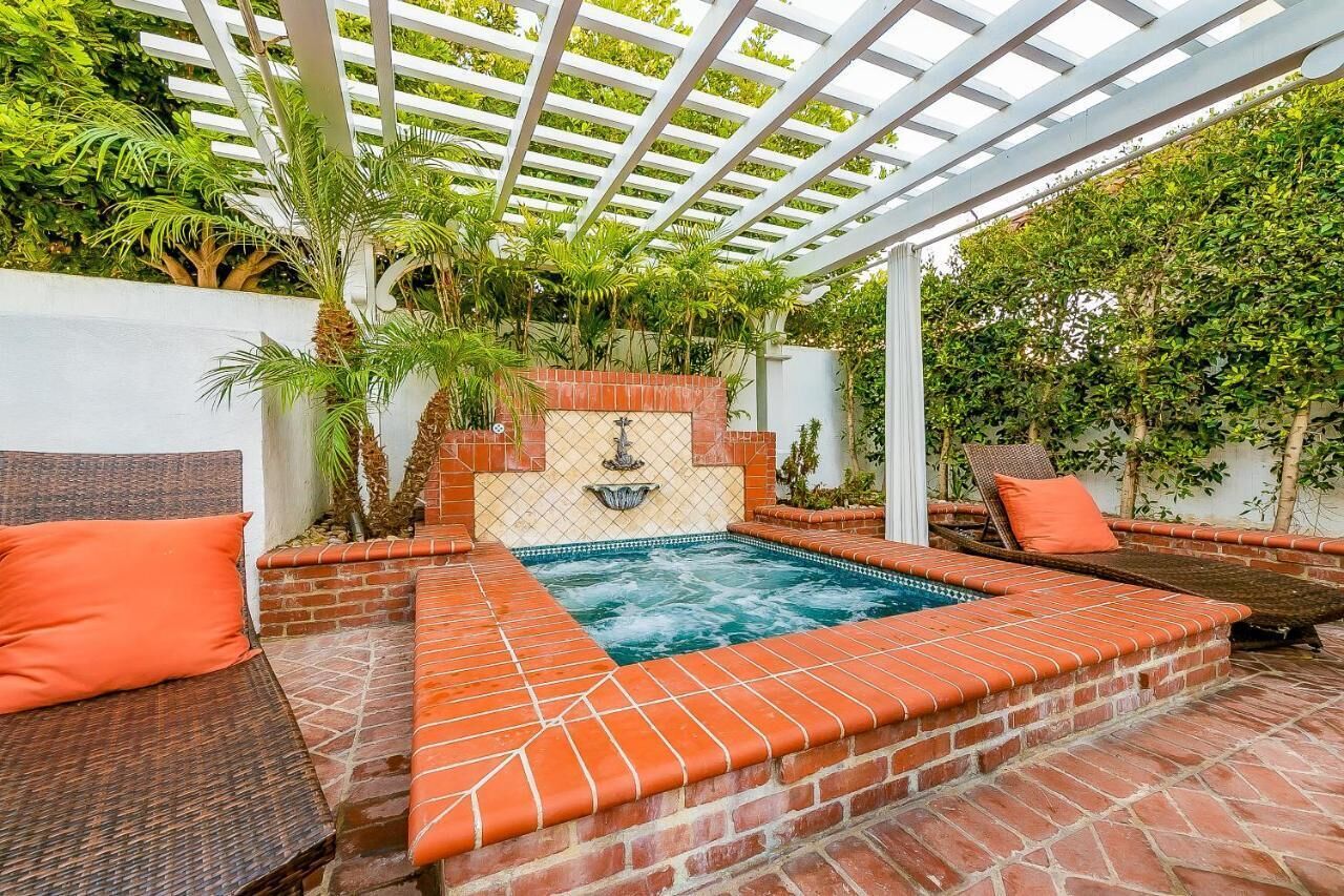 A covered outdoor swimming pool with a brick border, two lounge chairs, and a pergola overhead.