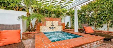 A covered outdoor swimming pool with a brick border, two lounge chairs, and a pergola overhead.
