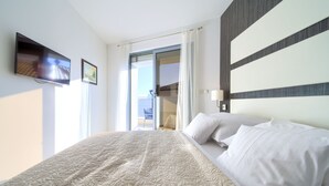Habitación de hotel con una cama grande, un televisor de pantalla plana montado en la pared, una puerta corrediza de vidrio al balcón y una lámpara moderna.