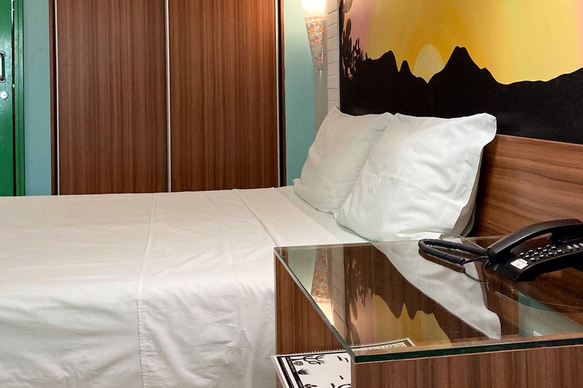 Ein Hotelzimmer mit einem Bett, einem Nachttisch mit Telefon, einer Wandlampe und einem Sonnenuntergangsbild.