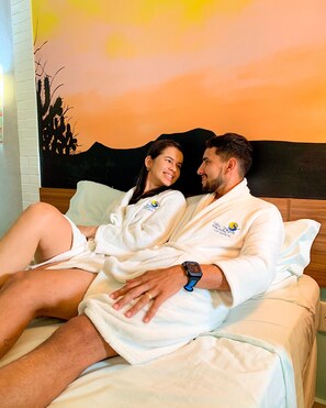 A couple in white bathrobes sitting on a bed with a painting of a sunset and mountains in the background. - Hotel e balneário olho d agua do milho (Caraúbas)