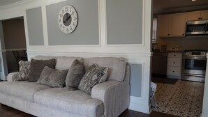 Un salón con un sofá gris, cojines estampados, un reloj de pared y una zona de cocina con armarios y electrodomésticos.