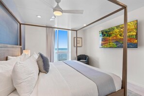 A hotel room with a four-poster bed, a ceiling fan, a large window with curtains, a chair, and a painting on the wall.