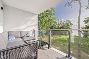 Ein Balkon mit Glastrennwand, ein graues Sofa mit Kissen und Blick auf eine Rasenfläche und Bäume.