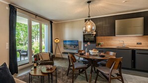 A modern living room with a sofa, dining table, and chairs, a kitchen with a large stove and cabinets, and a view through a sliding glass door. - Ferienwohnung Terrasse am Haff - Hochwertig Eingerichtetes Ferienappartment in Ruhiger Lage mit Wlan (Zirchow)