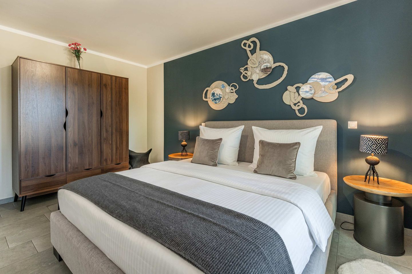 A modern bedroom with a large bed, a wooden wardrobe, decorative wall art, and two bedside tables with lamps.