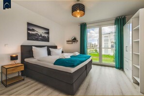 A bedroom with a bed, a nightstand, a lamp, a painting, a window with curtains, and a view of the outside.