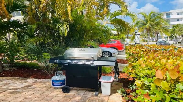 An outdoor grill setup with a propane tank, surrounded by lush greenery and palm trees.