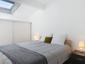 Un dormitorio con tragaluz, una cama con manta gris, dos lámparas de noche y un cabecero blanco.