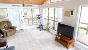 Ein geräumiges Wohnzimmer mit Deckenventilator, Laufband, Fernseher auf einem Holzboden und großen Fenstern mit Blick ins Freie.